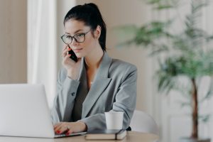 Serious brunette woman keyboards information, concentrated into laptop computer, dressed in formal outfit, poses at coworking space, works remotely, involved in working process during daytime.